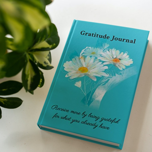 The hardcover version of the gratitude journal by Arisateam near a green plant.